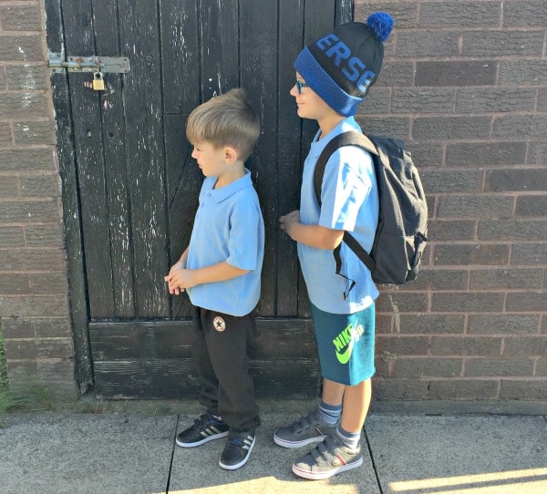 Two young boys wearing light blue polo shirts stand outside near a wooden door. Sporting P.E. Ready style from Get The Label, one has a rucksack, blue shorts, sunglasses, and a hat; the other wears black trousers. Both are facing left, ready for school.