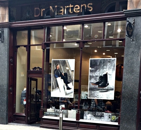 Shopfront of Dr. Martens with large glass windows displaying two posters: one of a person sitting in boots and another featuring a close-up of a black boot—perfect for your back to school style. The shop interior with shoes is visible inside.