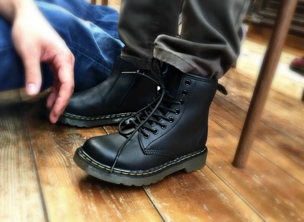 A person wearing classic Dr. Martens black lace-up boots with yellow stitching sits on a wooden floor, while another person in blue jeans, possibly ready for back to school, sits nearby, partially visible.