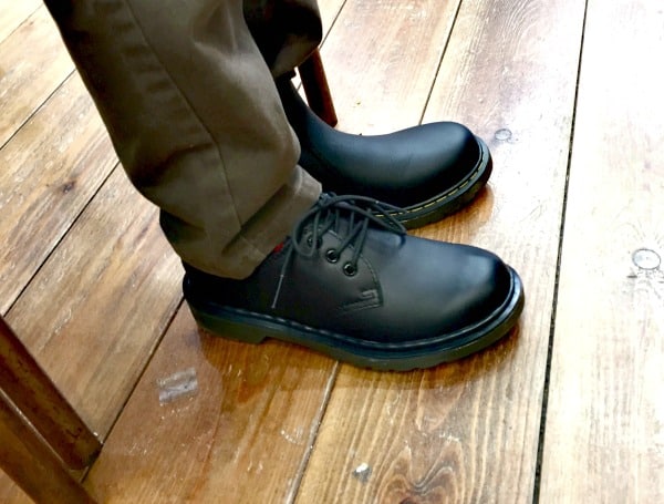 A person wearing black Dr. Martens lace-up shoes and brown trousers is sitting on a wooden chair, with their feet on a polished wooden floor—ready for back to school.