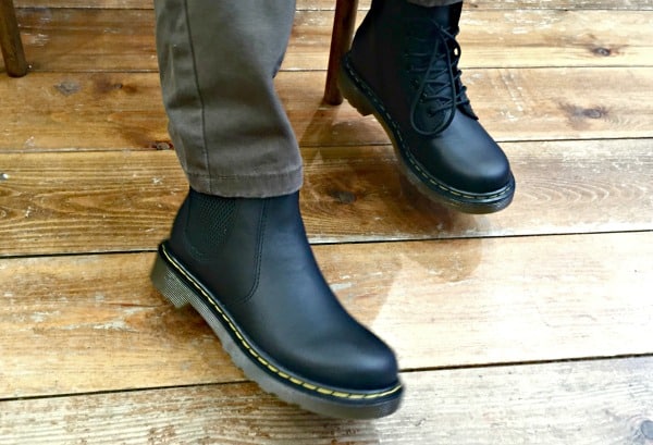 A person wearing brown trousers and black Dr. Martens boots stands on a wooden floor—one boot is lace-up, the other a Chelsea style—both featuring yellow stitching. Perfect for a fashionable back to school look.
