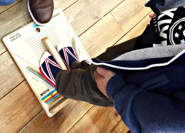 A child wearing Union Jack Dr. Martens stands on a shoe size measuring device, while an adult’s foot is visible nearby on a wooden floor—perfect for back to school preparations.