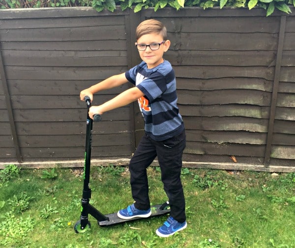 A young boy wearing glasses, a navy striped t-shirt, and black trousers stands on a fun two-wheeled black scooter on grass in front of a wooden fence, looking at the camera and smiling.