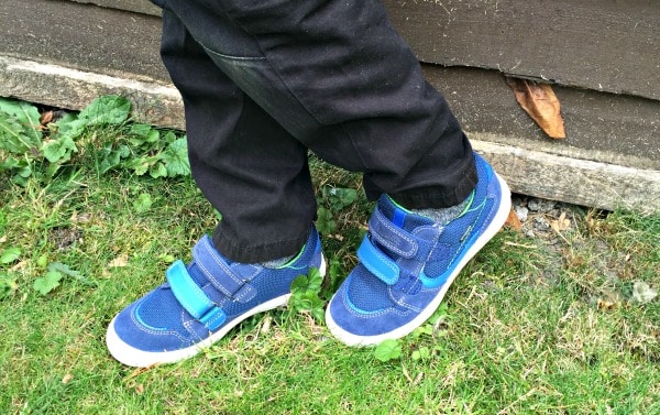 A child wearing blue trainers with velcro straps stands on grass with legs crossed, next to a wooden wall, ready for fun adventures on two wheels.