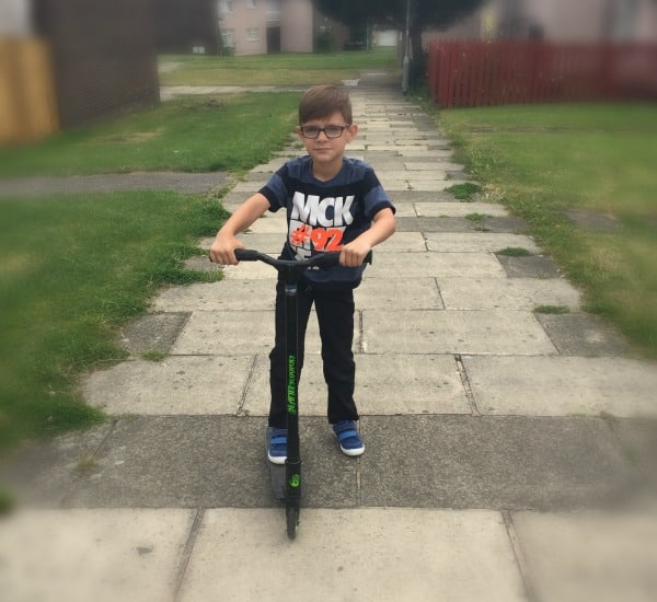 A young boy wearing glasses, a dark T-shirt, and trainers stands on a paved path holding the handlebars of a scooter, ready for fun on two wheels. Grass and houses are visible in the background.