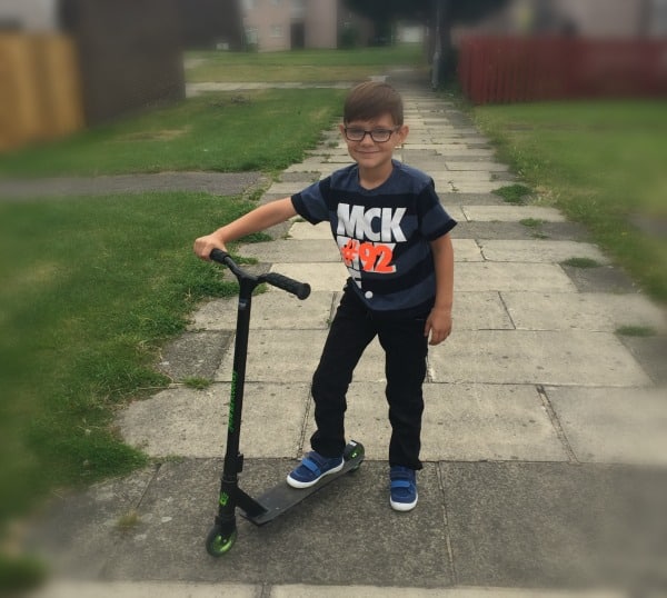 A young boy with glasses stands on a scooter, smiling as he enjoys fun on two wheels. He wears a striped T-shirt, dark trousers, and blue trainers. Grass and houses line the background along the paved pavement.