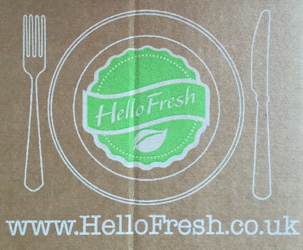 A cardboard surface with a white outline of a plate, fork, and knife features the green HelloFresh logo in the centre, proudly showcasing the hellofresh brand above the website "www.HelloFresh.co.uk" printed below.