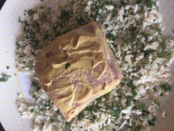A piece of raw meat coated with mustard rests on a plate with chopped green herbs and breadcrumbs, just like in a classic HelloFresh recipe, ready for breadcrumbing.