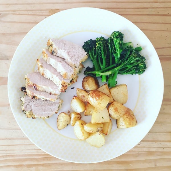 A white plate with sliced roast pork, sautéed tenderstem broccoli, and roasted potato pieces, in classic HelloFresh style, placed on a light wooden table.