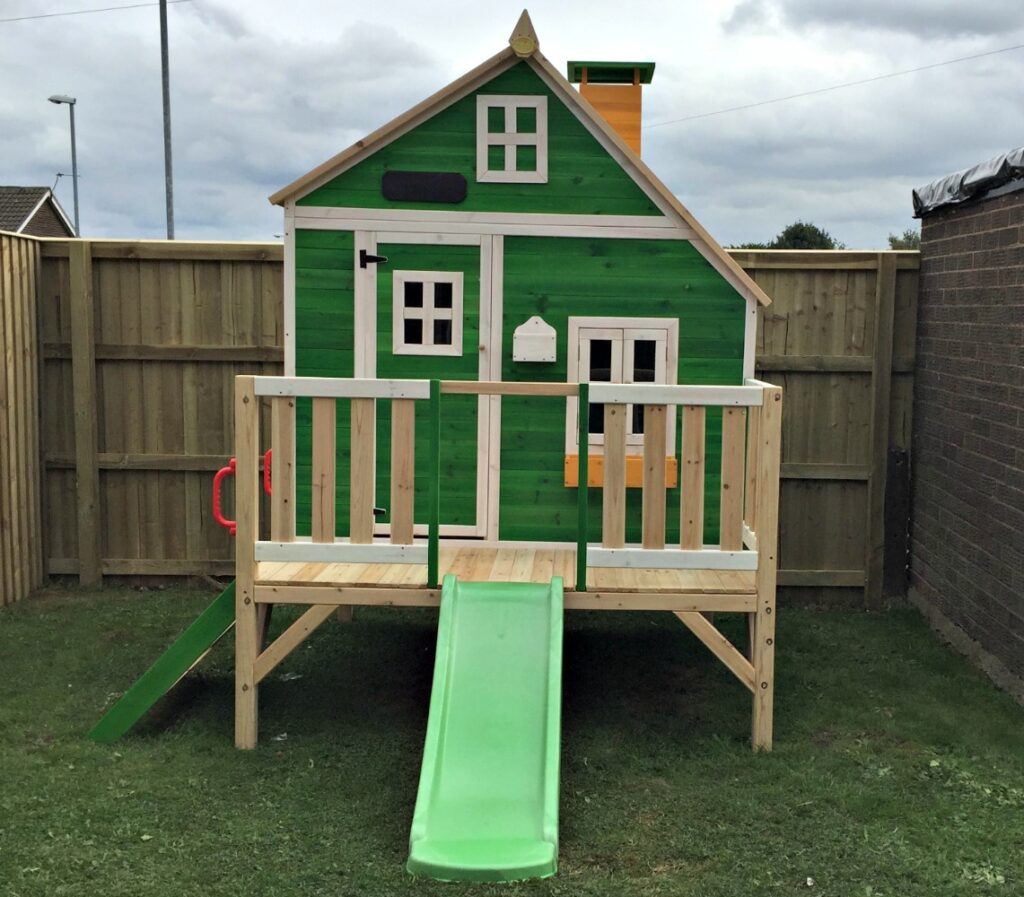 A small, green Wooden Playhouse by Big Game Hunters with white trim, a porch, windows, and a bright green slide sits on a grassy lawn, surrounded by a wooden fence and brick wall.