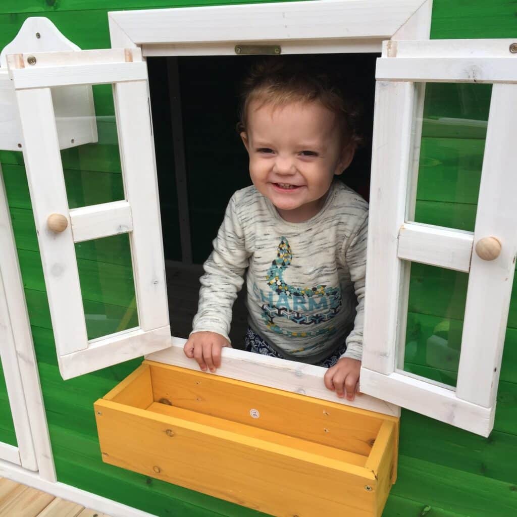 A smiling toddler stands at an open window of a Big Game Hunters Whacky Penthouse Wooden Playhouse with white trim and a yellow window box, looking outside.