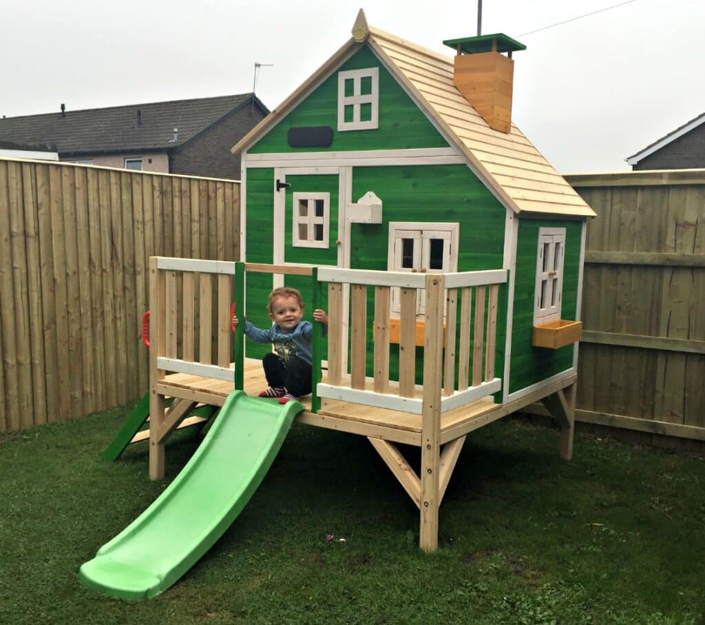 A young child sits on the porch of a Big Game Hunters Whacky Penthouse, a small green wooden playhouse with a slide in a fenced back garden. The elevated playhouse features white trim and a cheerful yellow chimney.