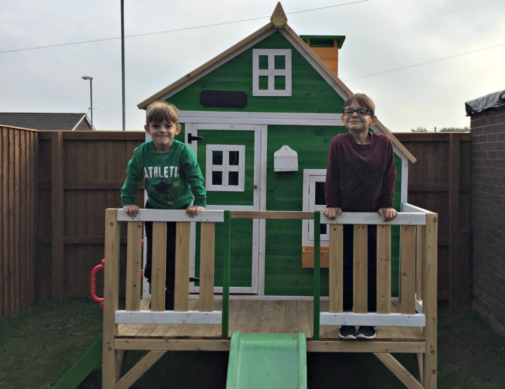 Two young boys stand on the porch of a green Big Game Hunters Penthouse Wooden Playhouse with a slide, in a fenced back garden. Both are smiling and holding the railing, with the playhouse behind them.