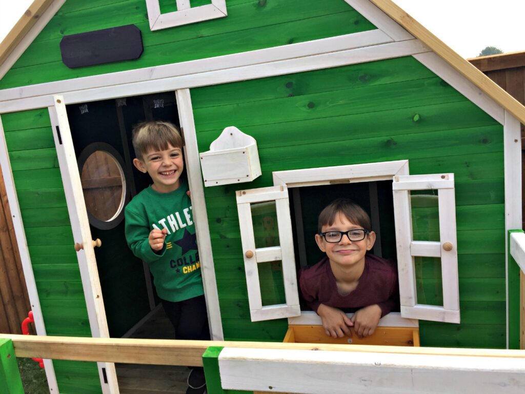 Two young boys smile and pose at a small green Whacky Playhouse by Big Game Hunters; one stands in the doorway while the other leans out of an open window. The house features crisp white trim and a fence in the foreground.