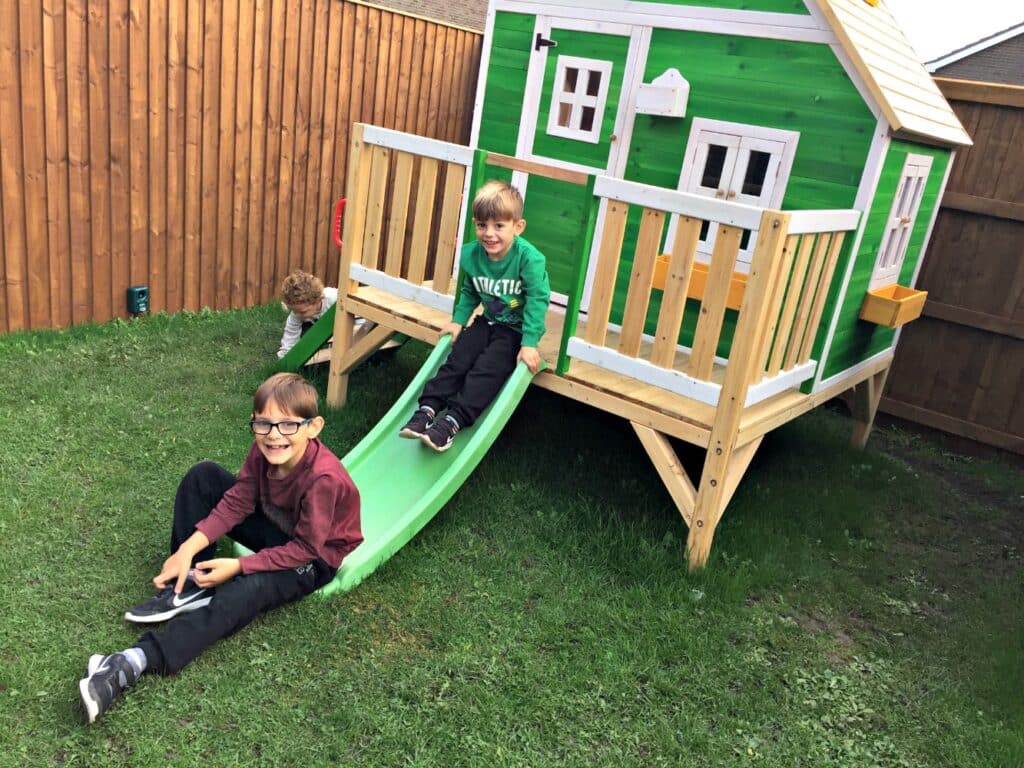 Three young boys play outside on a green slide attached to a green and white Penthouse Wooden Playhouse by Big Game Hunters. Two boys smile on the slide, whilst the third boy crouches on the grass nearby.