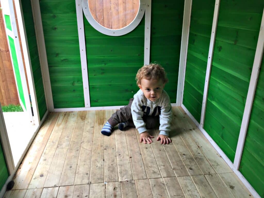 A toddler with curly hair crawls on the wooden floor inside a Big Game Hunters Wooden Playhouse with a round window, lit by sunlight streaming through the open door.