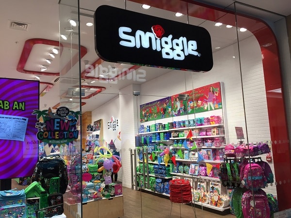 The entrance of a Smiggle shop featuring colourful stationery, bags, and rucksacks on display inside, with a large black and white Smiggle sign above the doorway, welcomes shoppers to the festive #SmiggleXmasParty.
