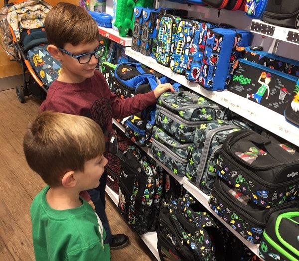 Two young boys look at a display of colourful lunch boxes on shop shelves at the #SmiggleXmasParty. One boy wears glasses and a maroon shirt, while the other has a green shirt. They appear to be deciding which lunch box to choose.
