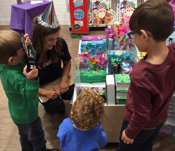 A woman in a party hat smiles whilst helping three young children choose toys from a colourful display at the #SmiggleXmasParty. The children look excited as they examine the festive items.