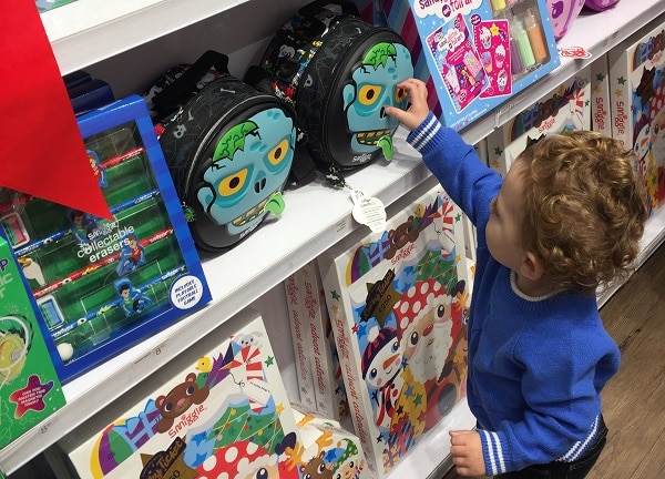 A young child in a blue jumper reaches for a monster-themed rucksack on a shop shelf filled with colourful toys and school supplies at the #SmiggleXmasParty.