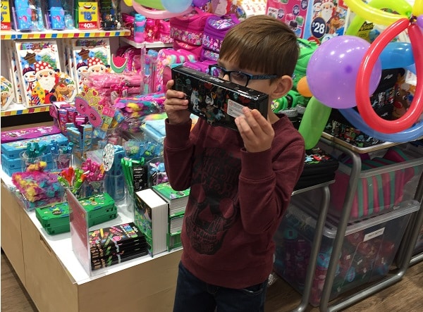 A young boy wearing glasses holds a toy box up to his face in a colourful toy shop filled with gifts, balloons, and various toys, ready for the #SmiggleXmasParty.