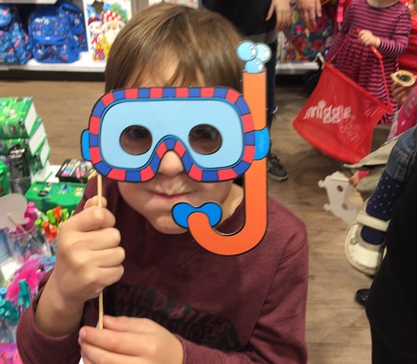 A child holds up a colourful cardboard cut-out of a snorkel and mask to their face at the #SmiggleXmasParty in a toy shop, with other children and toys visible in the background.