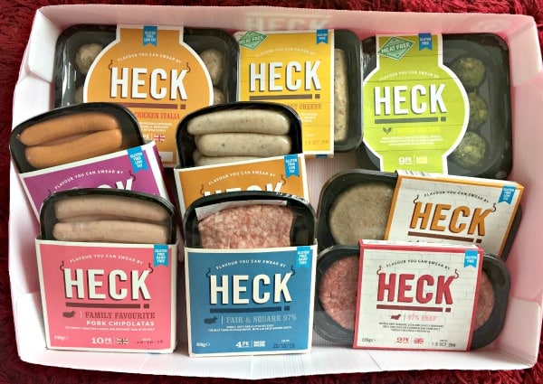 A selection of HECK sausage packs in various flavours and colours, including pork, chicken, and meat-free options, arranged as the Full Heck Deck in a box on a red surface.