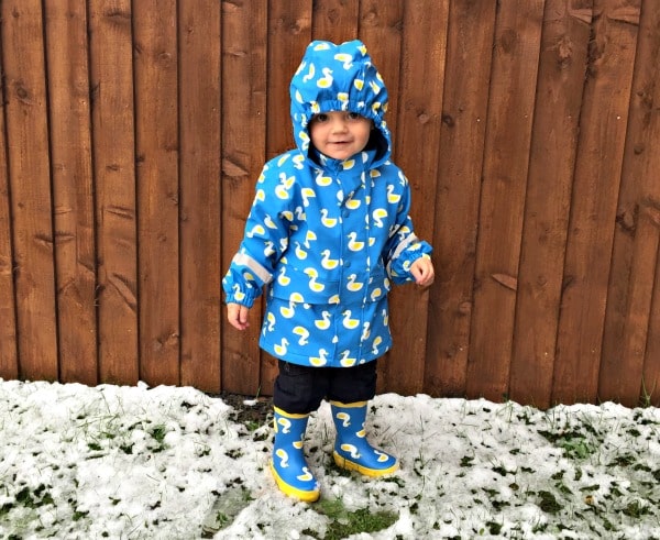 A young child stands on patchy snow in front of a wooden fence, wearing a blue raincoat and matching wellies decorated with yellow ducks, ready for snowy puddle adventures.