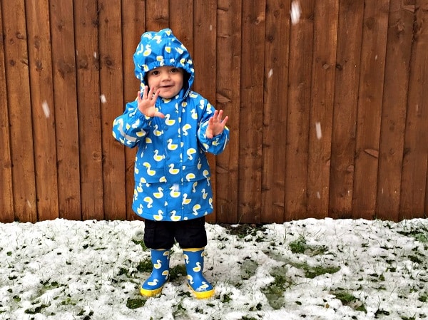A young child wearing a blue raincoat and wellies with yellow duck prints stands on patchy snow in front of a wooden fence, smiling and holding up one hand as snow falls—ready for more Snowy Puddle Adventures.