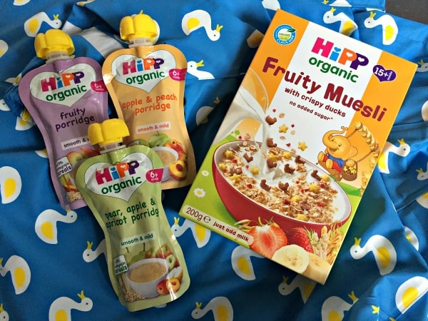 Three HiPP Organic baby food pouches and a box of HiPP Organic Fruity Muesli rest on a blue blanket with white and yellow duck prints, perfect for fuelling little ones before their next snowy puddle adventures.