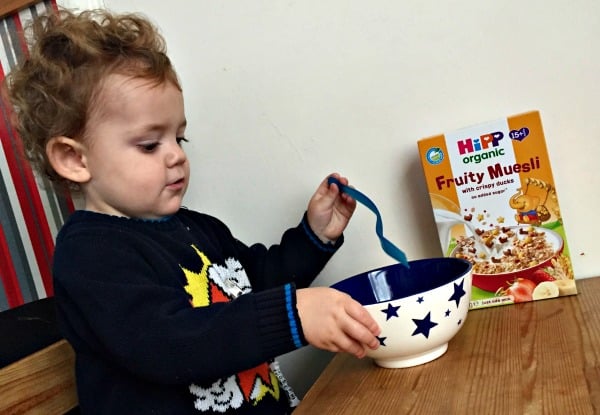 A young child sits at a wooden table, holding a spoon and a bowl with star patterns. A box of HiPP Organic Fruity Muesli cereal is on the table beside them, ready to fuel their next snowy puddle adventures.