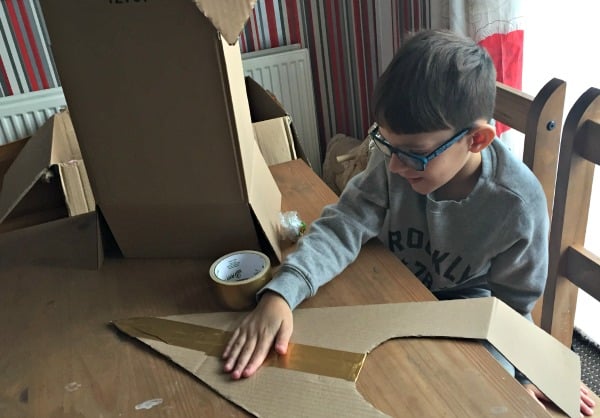 A boy wearing glasses sits at a wooden table, smoothing tape onto a piece of cardboard for a Family Challenge. Cardboard boxes, tape, and crafting materials are on the table, with striped wallpaper in the background.