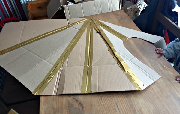 A large piece of card cut into a wing-like shape, with several strips of gold tape attached, is placed on a wooden table—perfect for an Energizer Family Challenge, with a child’s hand visible at the edge.