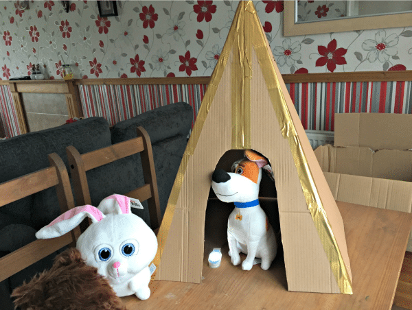 A stuffed bunny and a stuffed dog, inspired by The Secret Life of Pets, sit on a table. The dog is inside a cardboard tepee decorated with gold tape, while the bunny is just outside. The background shows floral wallpaper and a mirror.