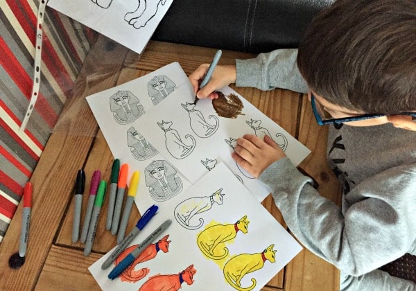 A child wearing glasses colours cat drawings on paper with felt-tip pens at a wooden table, inspired by The Secret Life of Pets. Several coloured and black-and-white cat illustrations and felt-tip pens are spread out on the table.