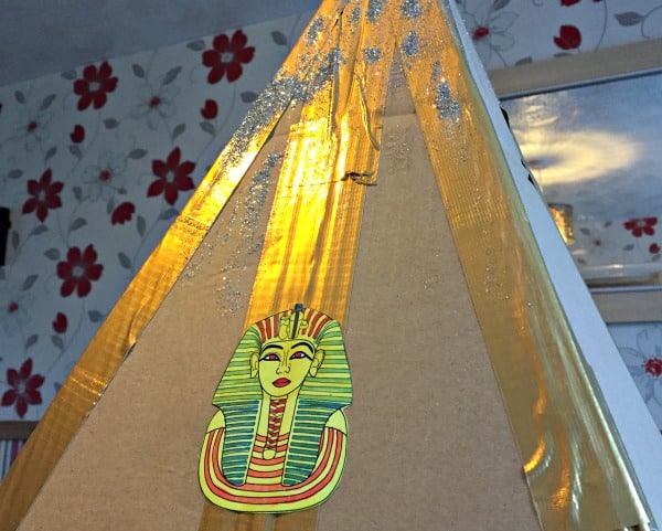 A cardboard pyramid decorated with gold tape and glitter, featuring a colourful drawing of an Egyptian pharaoh's mask—a perfect Family Challenge Energiser. A floral wallpaper and mirror are visible in the background.