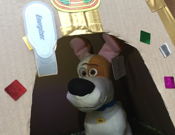 A plush toy dog sits inside a cardboard kennel decorated with colourful gem stickers and a gold ribbon, with a white Energizer clip light attached above the entrance, inspired by The Secret Life of Pets.