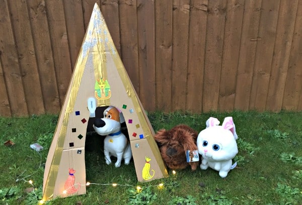 Three soft toy animals—a dog, a brown guinea pig, and a white cat from The Secret Life of Pets—stand beside a cardboard tepee decorated with colourful shapes and cat drawings, set on grass with a wooden fence in the background.