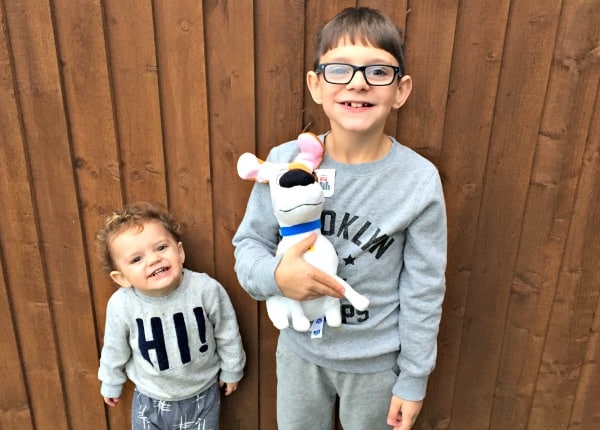 Two young children stand in front of a wooden fence. The older child, in glasses and a grey sweatshirt, holds a white plush dog toy from The Secret Life of Pets. The younger child, also in grey with "HI!" on their sweatshirt, smiles widely.
