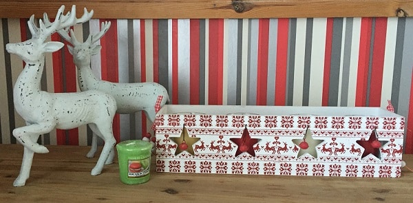 Two white decorative reindeer, a green candle, and a festive box with red and gold stars sit on a wooden surface in front of a red, grey, and white striped wall, creating charming Christmas décor for your home.
