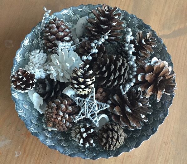 A decorative metal bowl filled with brown and white pine cones, silver beads, and a white star ornament makes charming Christmas home décor, beautifully displayed on a wooden surface.