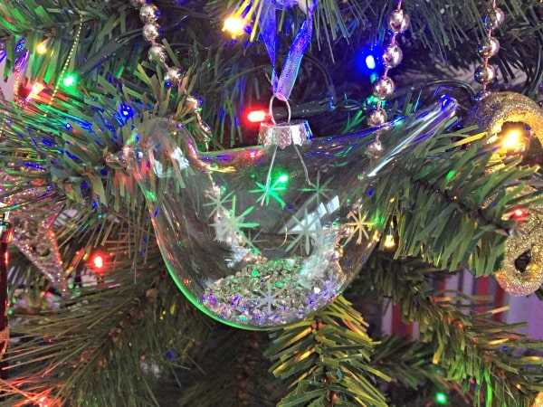 A clear glass bird ornament filled with silver glitter hangs on a Christmas tree, adding festive decor to the house and sparkling among multicoloured lights, tinsel, and other Christmas decorations.