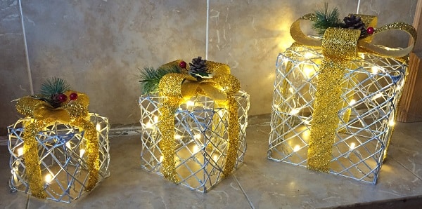 Three decorative gift boxes with gold ribbons and bows, lit from within by fairy lights, sit on a tiled surface. Perfect for Christmas home decor, each box is topped with festive greenery, red berries, and a pine cone.