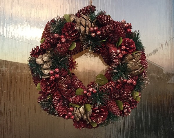 A festive Christmas wreath decorated with red pinecones, green artificial pine needles, gold-painted pinecones, red berries, and green leaves adds cheerful decor to a house’s textured glass door.