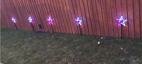 Five star-shaped decorative lights in red, white, and blue are lined up along a wooden fence in a grassy garden, adding festive charm to the house. A tennis ball and some small objects rest on the grass.