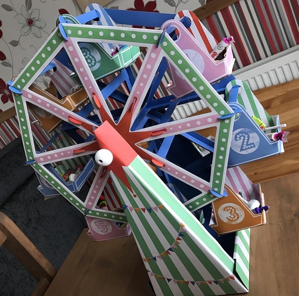 A colourful, striped cardboard Ferris wheel model from The Entertainer, with numbered pink, blue, and green cabins, sits on a wooden table—perfect for adding fun to family games or a Games Night In. The background features floral and striped wallpaper.