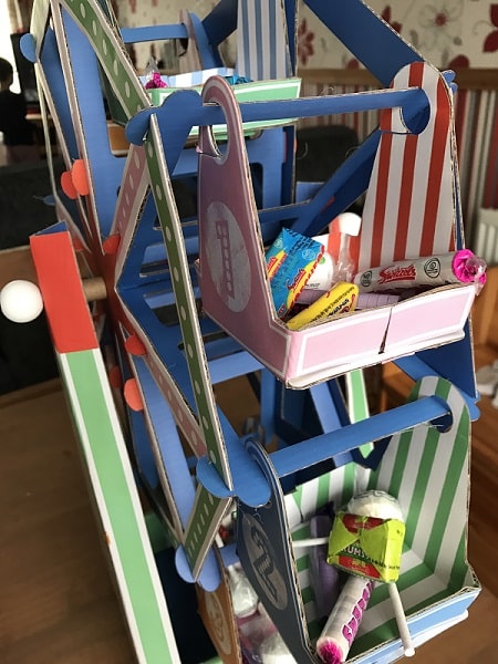 A colourful paper Ferris wheel from The Entertainer holds various sweets, including lollies and wrapped confectionery, in its gondolas. Perfect for a Games Night In, it sits on a wooden surface with striped and vibrant coloured sections indoors.