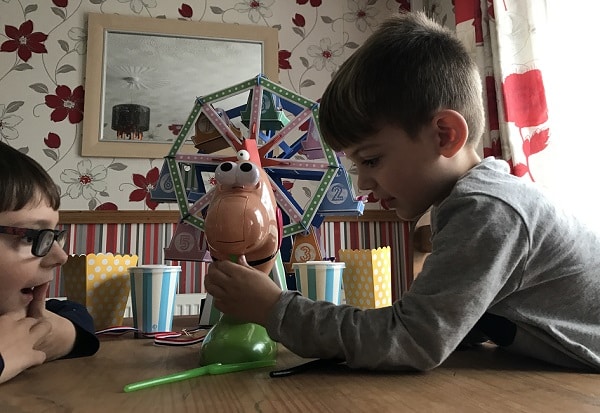 Two young boys enjoy a Games Night In, playing a tabletop game with a colourful Ferris wheel and cartoon face at a wooden table. Striped popcorn cups sit nearby, completing the fun scene in a room with floral wallpaper.