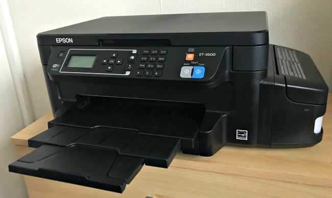 A black Epson EcoTank ET-3600 printer sits on a light-coloured wooden surface, its paper tray extended and control panel clearly visible on the front.