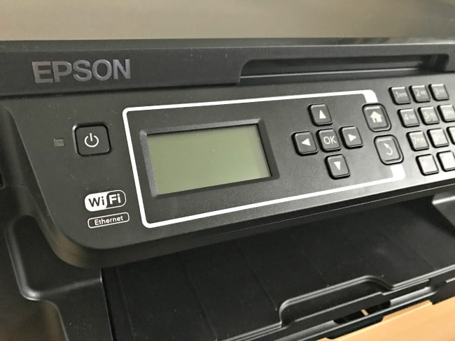 Close-up of an Epson EcoTank ET-3600 printer control panel showing buttons, a small display screen, and WiFi and Ethernet labels. The EcoTank printer tray below the panel is open.